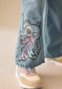 Picior de blugi cu personaj de desen animat și design cu text "AMCN", asortat cu adidași în culori pastel pe fundal deschis al podelei.