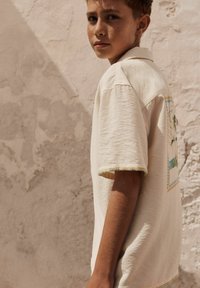Chemise beige à manches courtes avec un design brodé comprenant des accents verts et bleus, un tissu texturé et des coutures contrastées sur les manches.