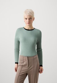 Vihreä ribattupinta pitkähihaine villapaita, jossa on musta reunus kaula-aukossa, yhdistettynä vaaleanbeigeihin korkeavyötäröisiin housuihin, joissa on sileä pinta ja istuva leikkaus.