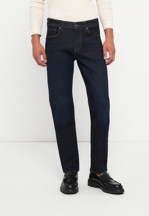 Dark blue denim jeans with a straight fit, front pockets, and a clean design. Worn with black shoes featuring a decorative strap.