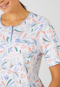 Camiseta de manga corta en tejido ligero con un patrón floral en tonos de naranja, verde y azul; incluye un escote abotonado y un bolsillo en el pecho.