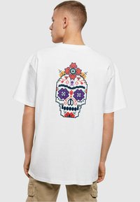 Hvit oversized t-skjorte med et fargerikt pikselert hodeskalle-design på ryggen, pyntet med blomster i rød, lilla og blå.