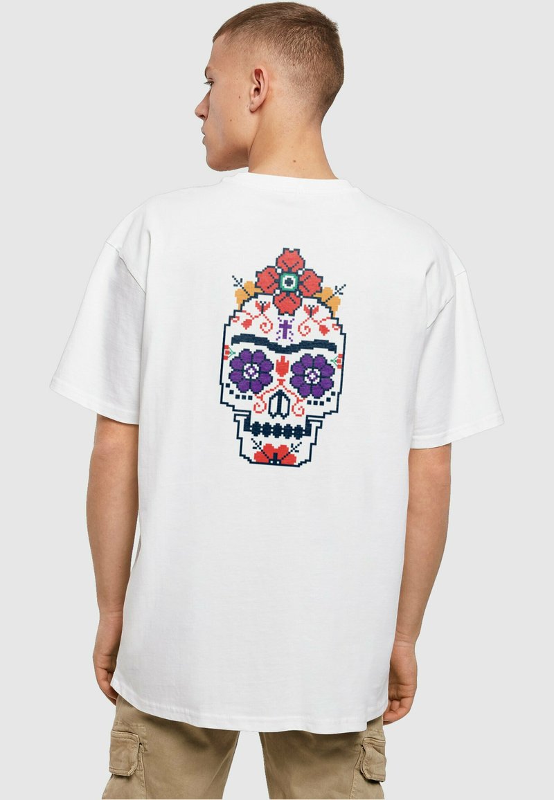 Hvit oversized t-skjorte med et fargerikt pikselert hodeskalle-design på ryggen, pyntet med blomster i rød, lilla og blå.