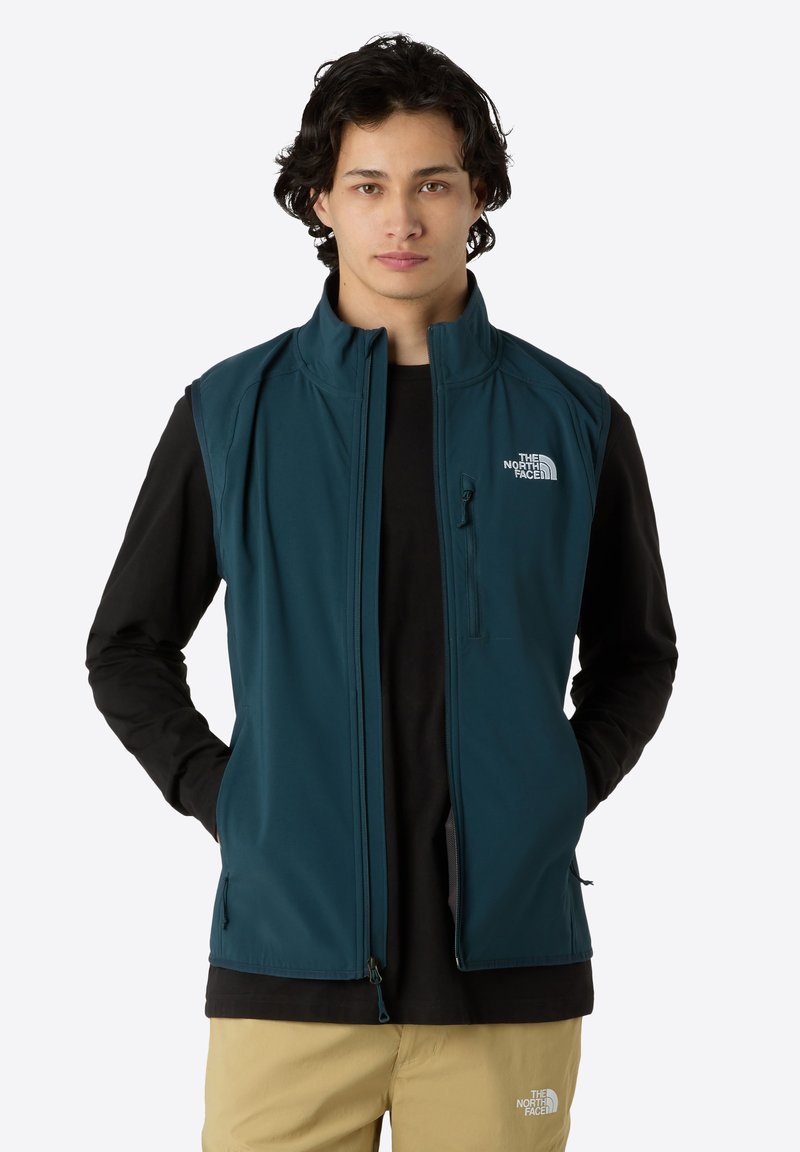 Tegelsvest met hoge hals, ritssluiting aan de voorkant en logo op de linkerkant van de borst; gecombineerd met een zwart shirt met lange mouwen en beige broek. Glad materiaal.