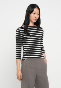 Stripete svart-hvit båt-hals genser, tre fjerdedeler ermer, myk stoff, med en liten logo detalj på brystet, kombinert med grå bukser.