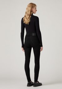 Šviesiaplaukė moteris su banguotais plaukais, vilkinti juodą raštuotą ilgam rankovėm megztinį, juodas siauras kelnes ir juodas loafers tipo batus, stovinti pasisukusi šonu.