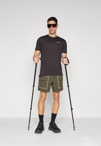 Μαύρο μπλουζάκι, ελαιόχρωμα cargo σορτς με διακοσμητικές τσέπες, μαύρες κάλτσες και παπούτσια, κρατώντας μπαστούνια πεζοπορίας. Το μοντέλο στέκεται μπροστά σε λευκό φόντο.