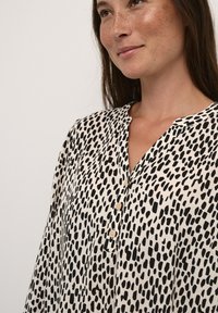 Koszula zapinana na guziki w kolorze kości słoniowej z czarnym wzorem w kropki, wykonana z lekkiego materiału, z dekoltem w kształcie litery V oraz trzema guzikami z przodu, o luźnym kroju.