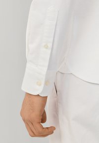 Mands venstre hånd ved siden af hvid skjortes manchet med to knapper, kombineret med lyse bukser.