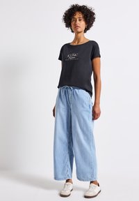 Černé tričko s krátkým rukávem s nápisem "ICONIC" na přední straně, doplněné o světle modré široké džíny a bílé tenisky.