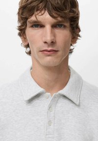 Szara koszulka polo z kołnierzykiem na zakładkę i guzikiem z przodu. Materiał wydaje się miękki, o subtelnej fakturze. Brak widocznych wzorów lub ozdób.