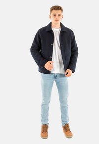 Giacca blu navy in misto lana con chiusura frontale con bottoni, due tasche e collo a rever, abbinata a jeans chiari e stivali marroni con lacci.