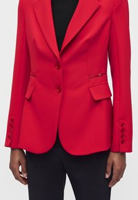 Blazer rouge sur mesure avec deux boutons, poches à rabat et détails de boutons aux manches, porté sur un top noir et un pantalon.