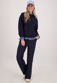 Marine sweatshirt over een blauwe denimshirt, gecombineerd met marinebroeken en witte sneakers. Geaccentueerd met een lichtblauwe pet met rode borduursels.