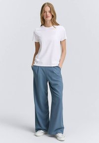 Bela majica z okrolim izrezom v kombinaciji z ohlapnimi, teksturiranimi svetlo modrimi hlačami. Model stoji z rokami v žepih in nosi bele superge.