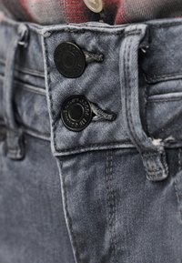 Κοντινό πλάνο της μέσης ενός γκρι τζιν με δύο μαύρα μεταλλικά κουμπιά και ένα κόκκινο πουκάμισο σε καρό, που είναι μπουφάν μέσα στη μέση.