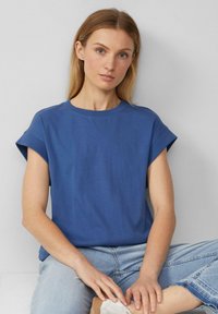 Blaues Baumwoll-T-Shirt mit kurzen Ärmeln und Rundhalsausschnitt, das eine lockere Passform und eine glatte Textur aufweist. Kombiniert mit hellblauen Jeans und weißen Schuhen.