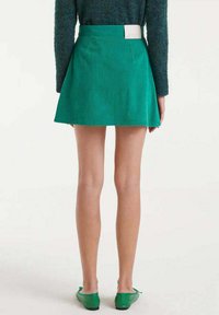 Mujer vestida con una mini falda de pana verde, una blusa de manga larga oscura y unos zapatos planos verdes, vista desde atrás sobre un fondo claro y liso.