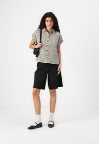 Camicetta a maniche corte bianca e nera a pois, abbinata a pantaloni corti neri su misura, scarpe basse nere con dettagli e calze grigie.