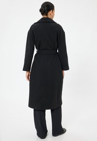 Cappotto trench in lana nera con maniche lunghe, vita con cintura e dettaglio sullo yoke posteriore. Presenta una vestibilità dritta e arriva a metà polpaccio.