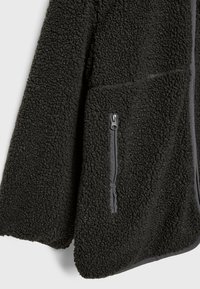 Zwart fleece jack met een gestructureerd, pluche oppervlak. Beschikt over een zijzak met een elegante rits. Afgewerkt met contrasterende bies langs de randen.