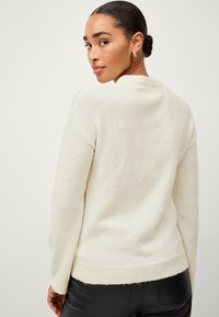 Suéter de punto color crema con dobladillo acanalado, que presenta un acabado texturizado; tiene un escote redondo y mangas largas, combinado con pantalones negros.