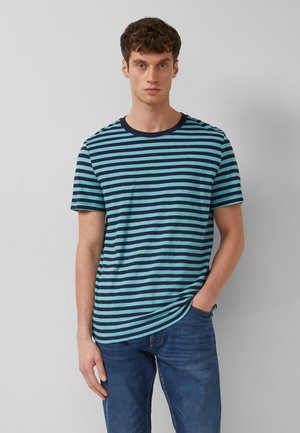 Homme portant un t-shirt à manches courtes à rayures horizontales bleu marine et bleu clair, ainsi qu'un jean bleu, debout devant un fond gris uni.