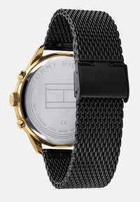 Montre en acier inoxydable doré avec un bracelet en maille tissée noire. Elle présente un cadran rond avec un fond transparent et une fermeture à clapet.
