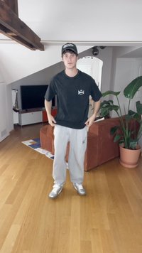 Fekete pamut póló logóval, szürke melegítőnadrág és ezüst sportcipő. Belső tér fából készült padlóval és cserépben lévő növénnyel.