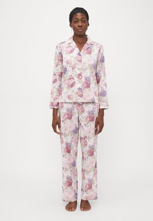 Kvinne som står barføtt iført en matchende pyjamas med blomsterprint, langermet knapp-up topp og bukser i rosa, lilla og beige nyanser.
