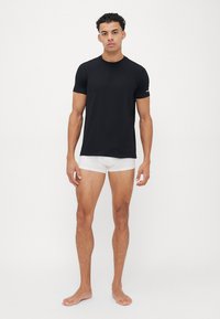 Schwarzes kurzärmliges T-Shirt aus glattem Stoff, taillierte Passform. Getragen mit weißen Unterhosen. Model steht barfuß vor einem schlichten Hintergrund.