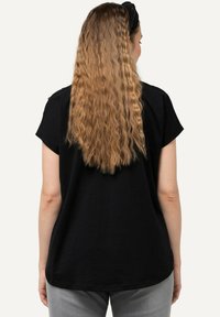 Top negro de manga corta con dobladillo redondeado y textura suave, con un ajuste relajado. El cabello es largo y ondulado, sin adornos visibles.