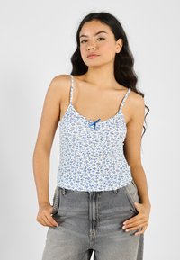 Hvit tanktop med blått blomstermønster, justerbare stropper og en liten blå sløyfe som aksent ved utringningen. Bært med grå høytlivsbukser.