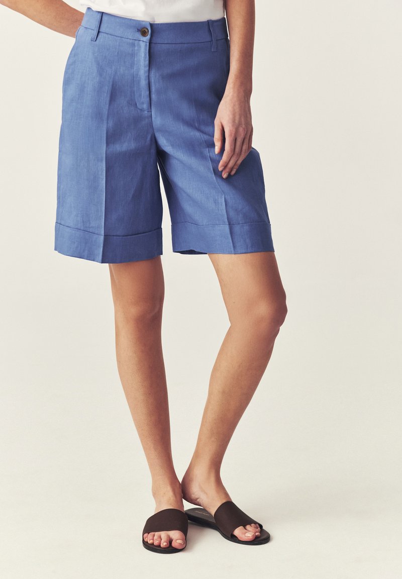 Mannequin portant un short bleu taille haute jusqu'aux genoux avec des revers et des sandales noires à bouts ouverts, debout devant un fond uni.