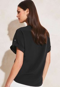 Blusa nera a maniche corte con risvolti e dettagli di bottoni lucidi, caratterizzata da una texture morbida e un orlo dritto.