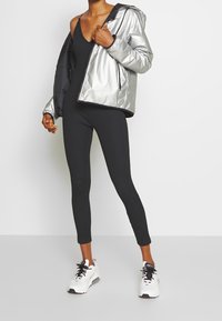 Černý body s metalickou stříbrnou zimní bundou s kapucí. Černé legíny a bílé sportovní boty s černými akcenty dokončují outfit.