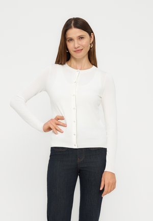 Kvinde med lige brunt hår iført gyldne hoops-øreringe, en hvid knappet cardigan og mørkeblå jeans, stående med den ene hånd på hoften.