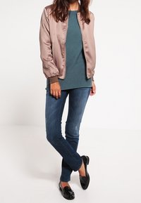 Lichtbruine satijnen bomberjack, teal gekleurde top met lange mouwen, donkerblauwe spijkerbroek en zwarte loafers. Model staat met één voet voor.