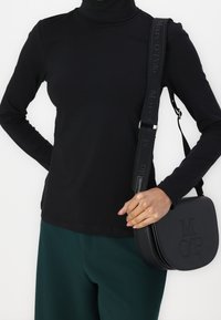 Black turtleneck top paired with dark green pants. A black crossbody bag with embossed "MOP" logo and a textured strap.