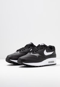 Les baskets Nike Air Max 1 noires présentent un dessus en cuir lisse, des accents blancs, des lacets ronds et une unité air visible dans la semelle. Semelle extérieure en caoutchouc texturé.