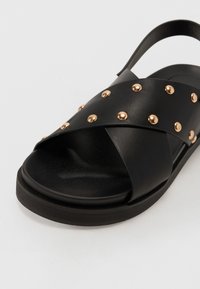 Svart sandaler med korskorsade remmar, prydda med metalliska guldstift. Slät textur och platt sula. Justerbar hälrem för passform.