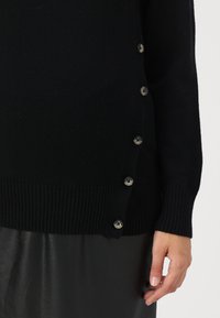 Svart strikket genser med ribbestrikkede mansjetter og hette, med dekorative knapper langs siden. Myk tekstur, minimalistisk design.