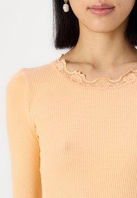 Katoenen geribbelde top in zachte perzik met een scalloped kant halslijn detail. De stof heeft verticale lijnen voor textuur.