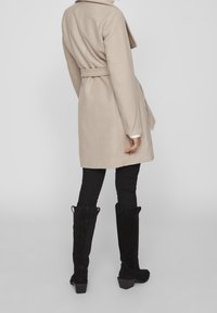 Beige kappa med bälte, krage, långa ärmar och sidofickor, bärs över svarta skinny byxor och svarta knähöga stövlar, som visar en mjuk textur.