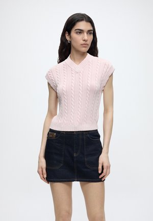 Kvinde iført en lyserød ærmeløs strikket sweater og en mørk denim mininederdel, stående mod en ensfarvet lys baggrund.