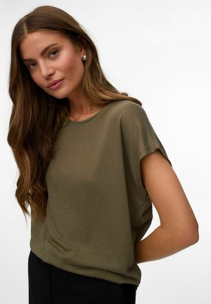 Jonge vrouw met lang bruin haar, gekleed in een olijfgroen kort mouwtopje en zilveren oorringen, staand tegen een effen witte achtergrond.