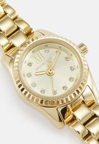 Montre-bracelet dorée avec un bracelet métallique. Le boîtier rond présente une lunette texturée, un cadran doré et des index horaires incrustés, avec le nom de la marque affiché.