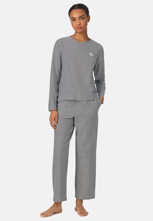 Vrouw staat blootsvoets, draagt een zwart-wit gestreept shirt met lange mouwen en bijpassende losse broek, hand in de zak, neutrale achtergrond.