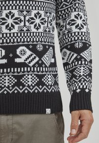 Strickpullover mit schwarz-weißen Schneeflockenmuster, gerippten Bündchen und Saum, mit einem kleinen Etikett am unteren Rand.
