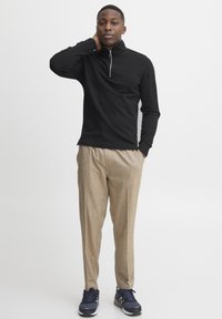Black zip-up long-sleeve top paired with light beige trousers and navy sneakers. Features a high collar and a zip closure at the neck.
